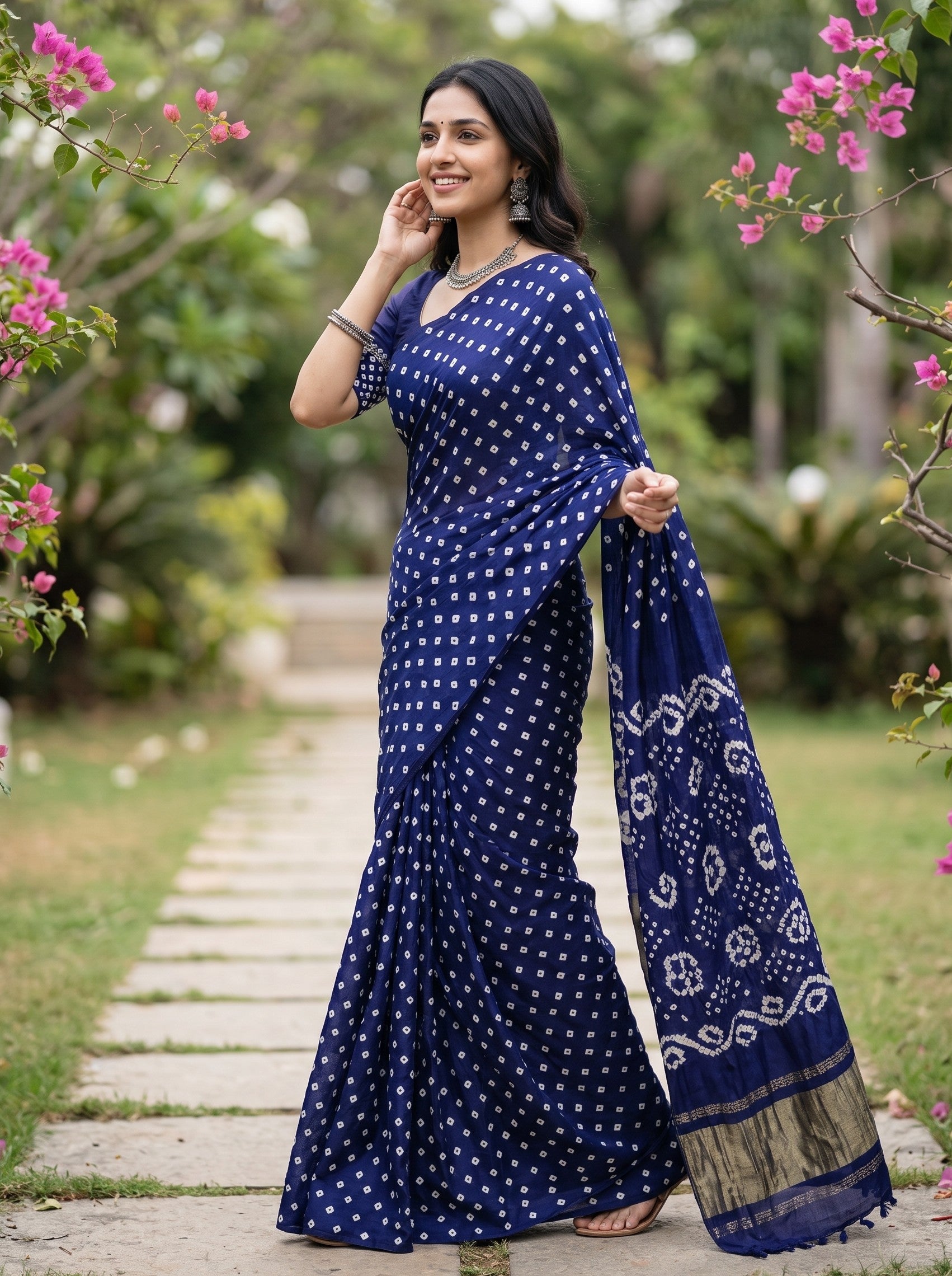 A woman wearing a navy blue saree with white tie-dye patterns and a gold border, standing on a stone path in a garden with pink flowers.