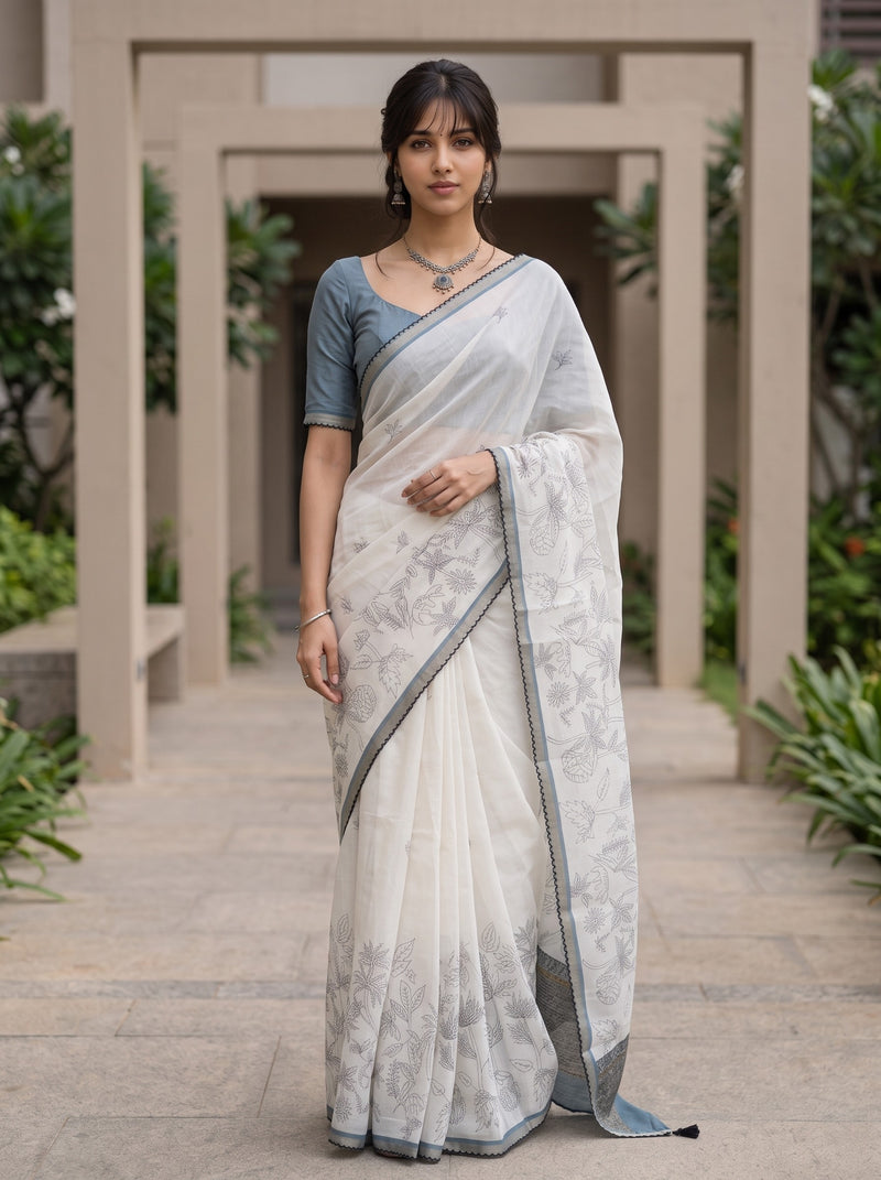 A woman wearing a white and gray patterned saree with a blue-gray short-sleeve blouse, standing in a garden corridor.