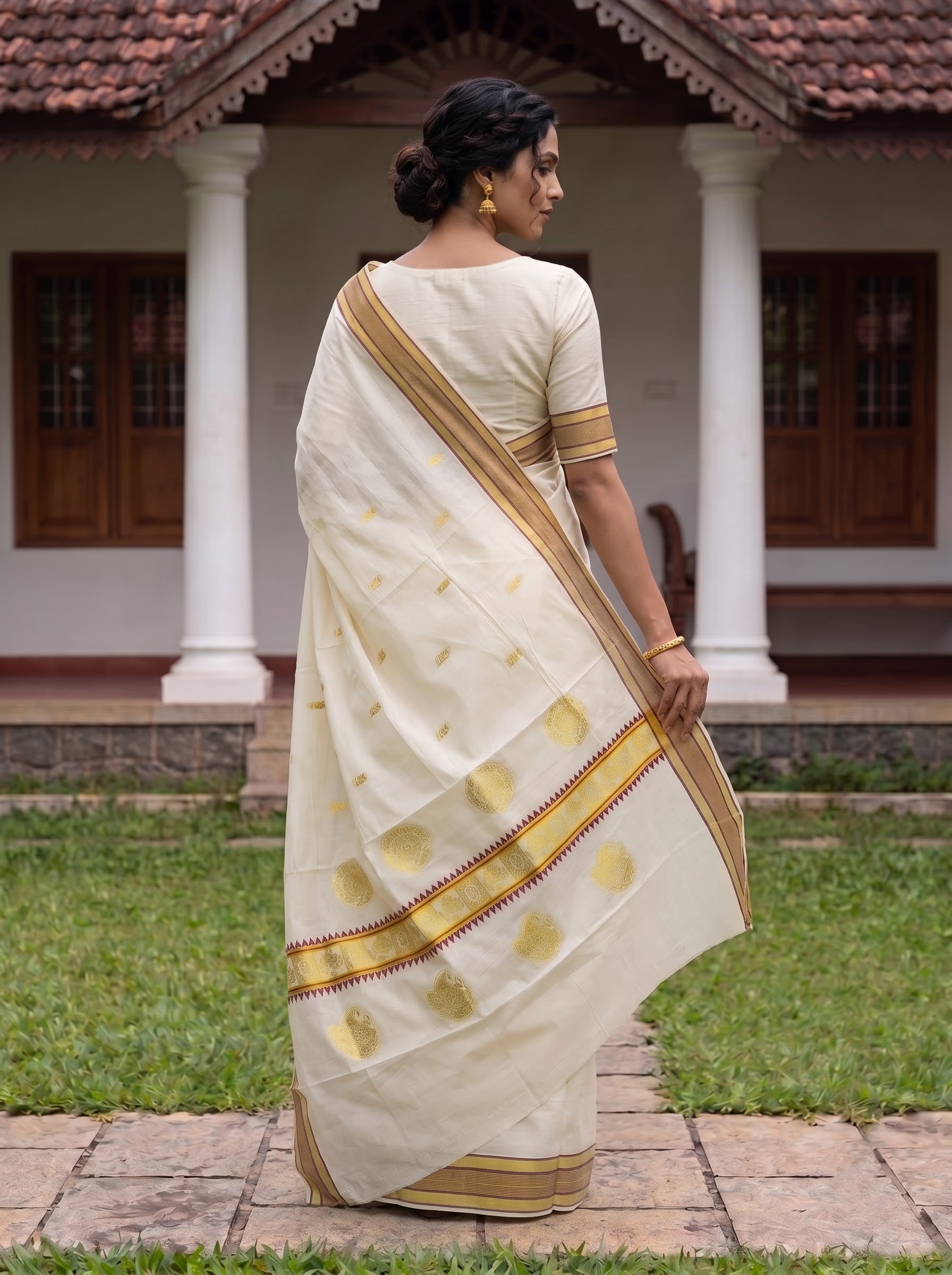 A woman wearing a traditional Indian saree stands outdoors in front of a house with white pillars and a tiled roof. The saree is off-white/cream with gold foil motifs and borders, paired with a cream blouse featuring gold trim.