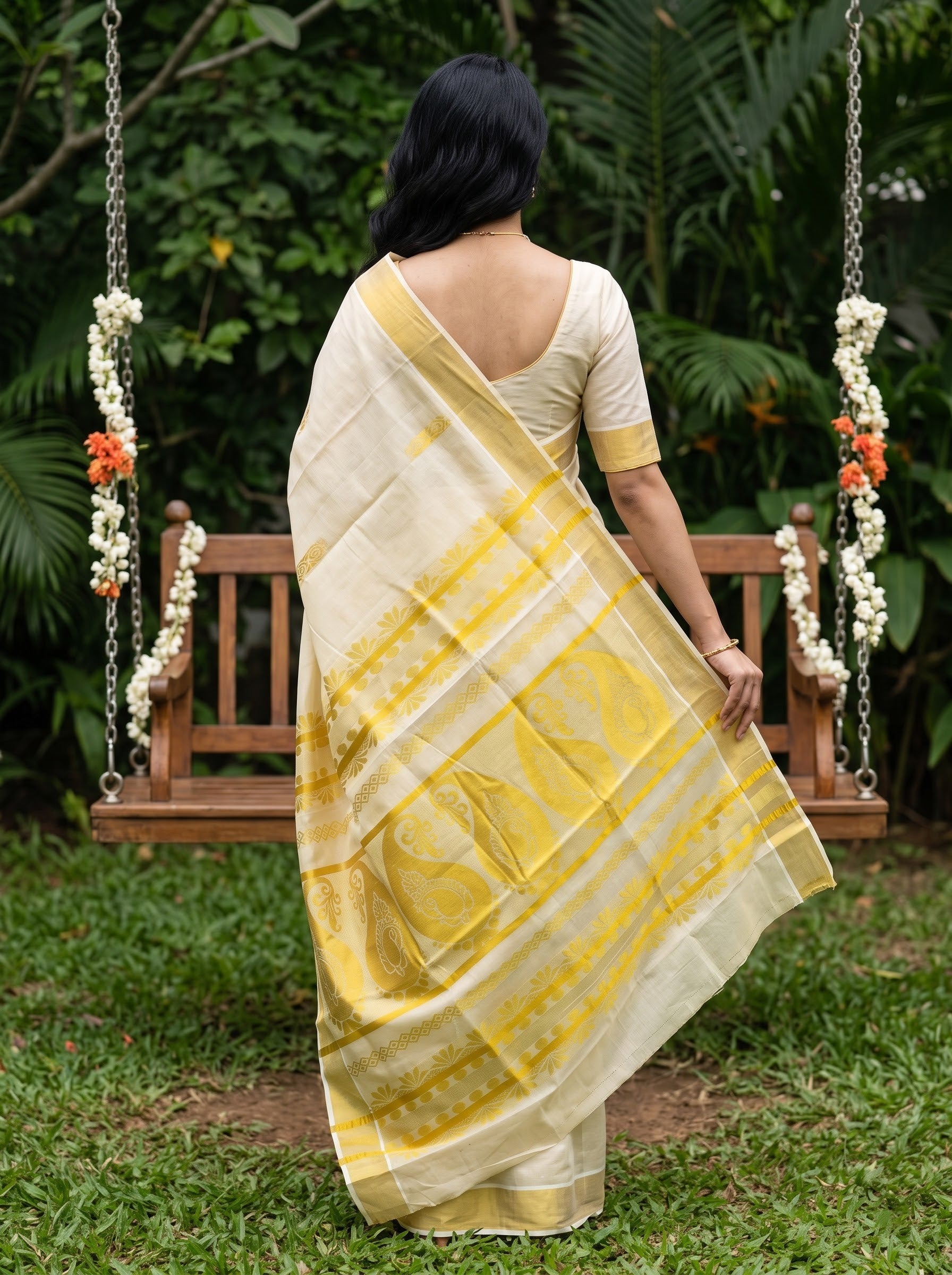 A woman stands with her back to the camera wearing a cream and yellow saree with a yellow printed blouse, posed in front of a wooden swing decorated with white and orange flower garlands in a lush green garden.