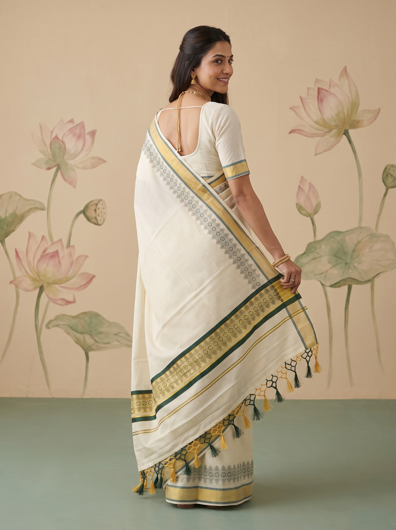 A woman modeling a white saree with green and gold border patterns, shown from the back against a beige wall with a painted lotus motif.