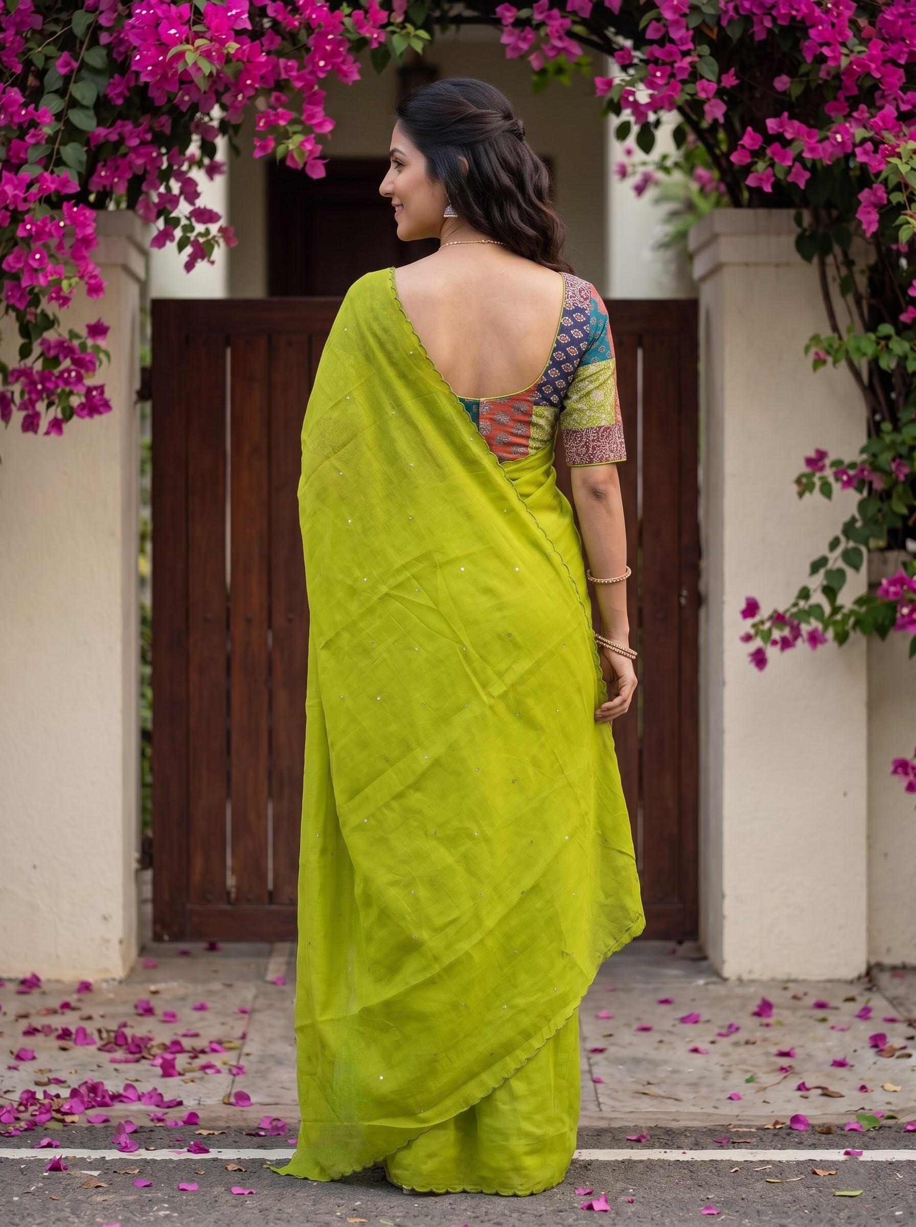 A woman wearing a bright lime green saree with a multicolored patterned blouse, standing in front of a wooden gate framed by pink bougainvillea flowers.