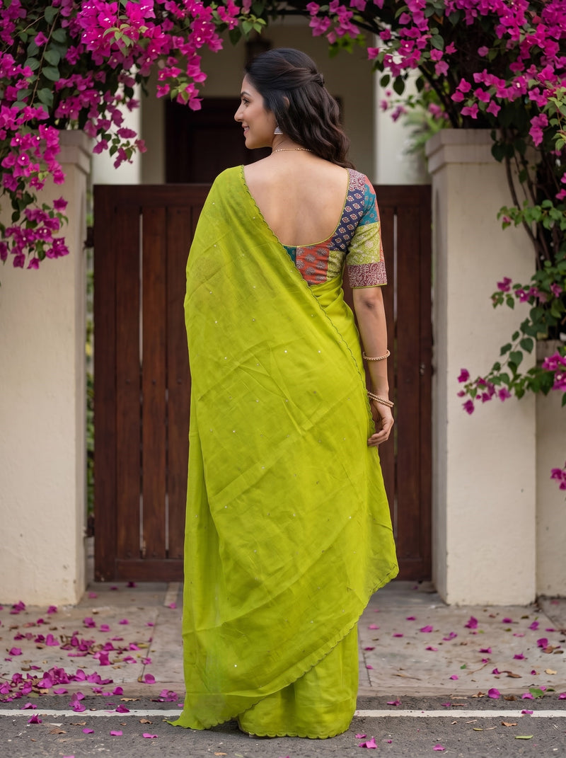A woman wearing a bright lime green saree with a multicolored patterned blouse, standing in front of a wooden gate framed by pink bougainvillea flowers.