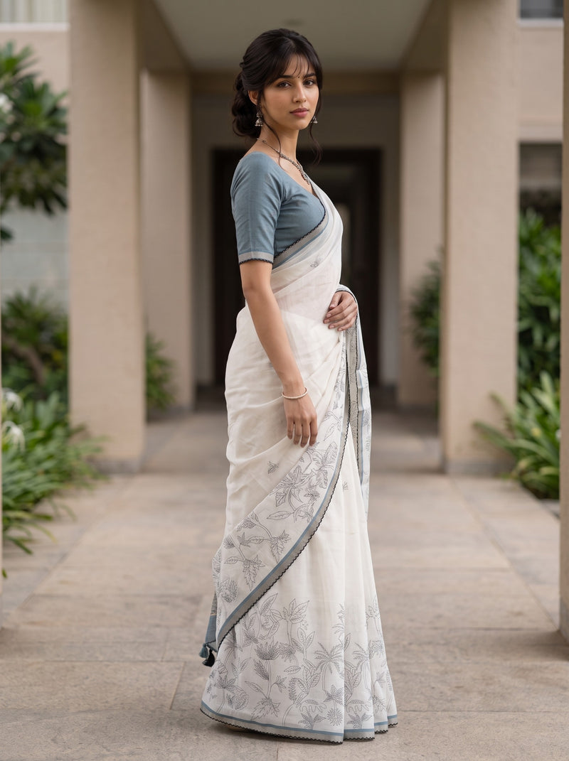 A woman wearing a white saree with gray floral patterns and a slate blue short-sleeve blouse, standing in a covered walkway.