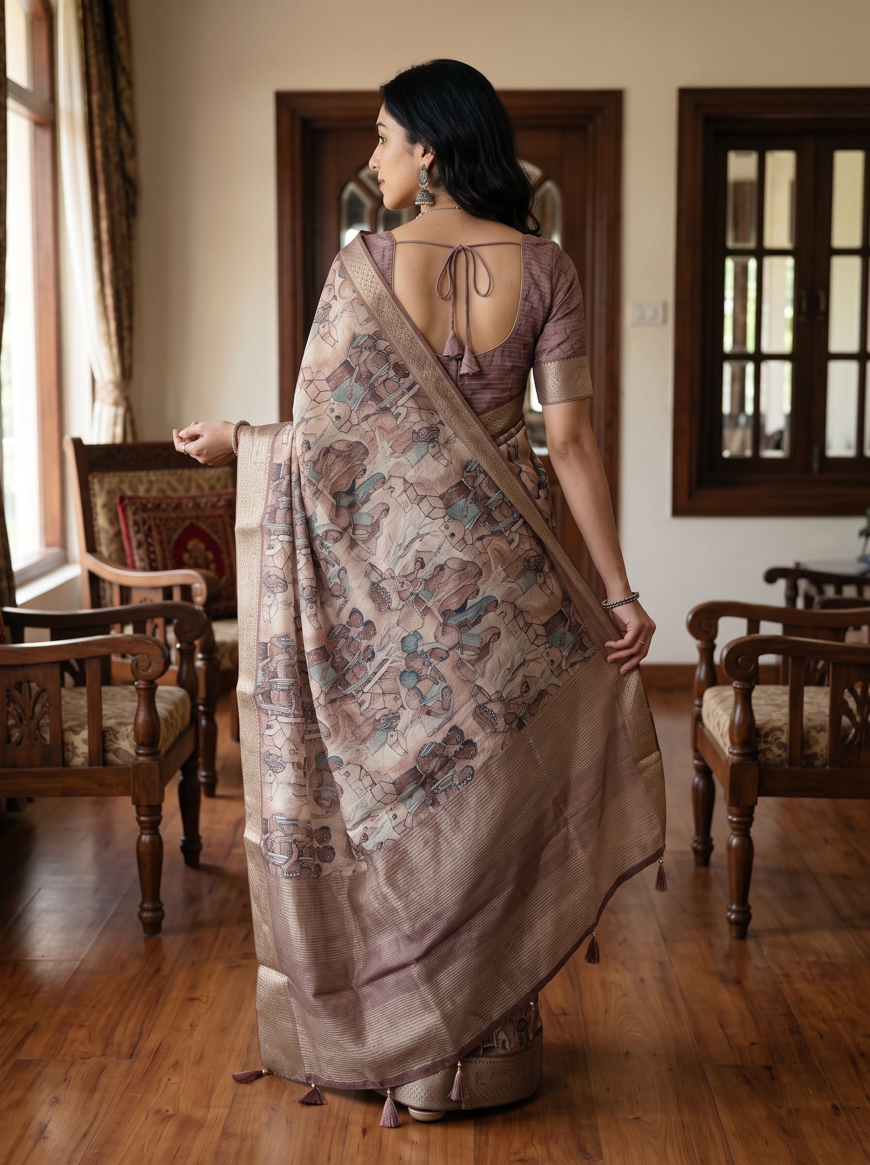 A woman stands in a room with wooden floors and chairs, wearing a draped saree with a muted mauve blouse and a patterned beige saree with blue and brown motifs.
