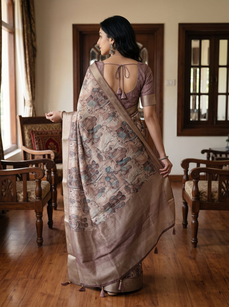A woman stands in a room with wooden floors and chairs, wearing a draped saree with a muted mauve blouse and a patterned beige saree with blue and brown motifs.