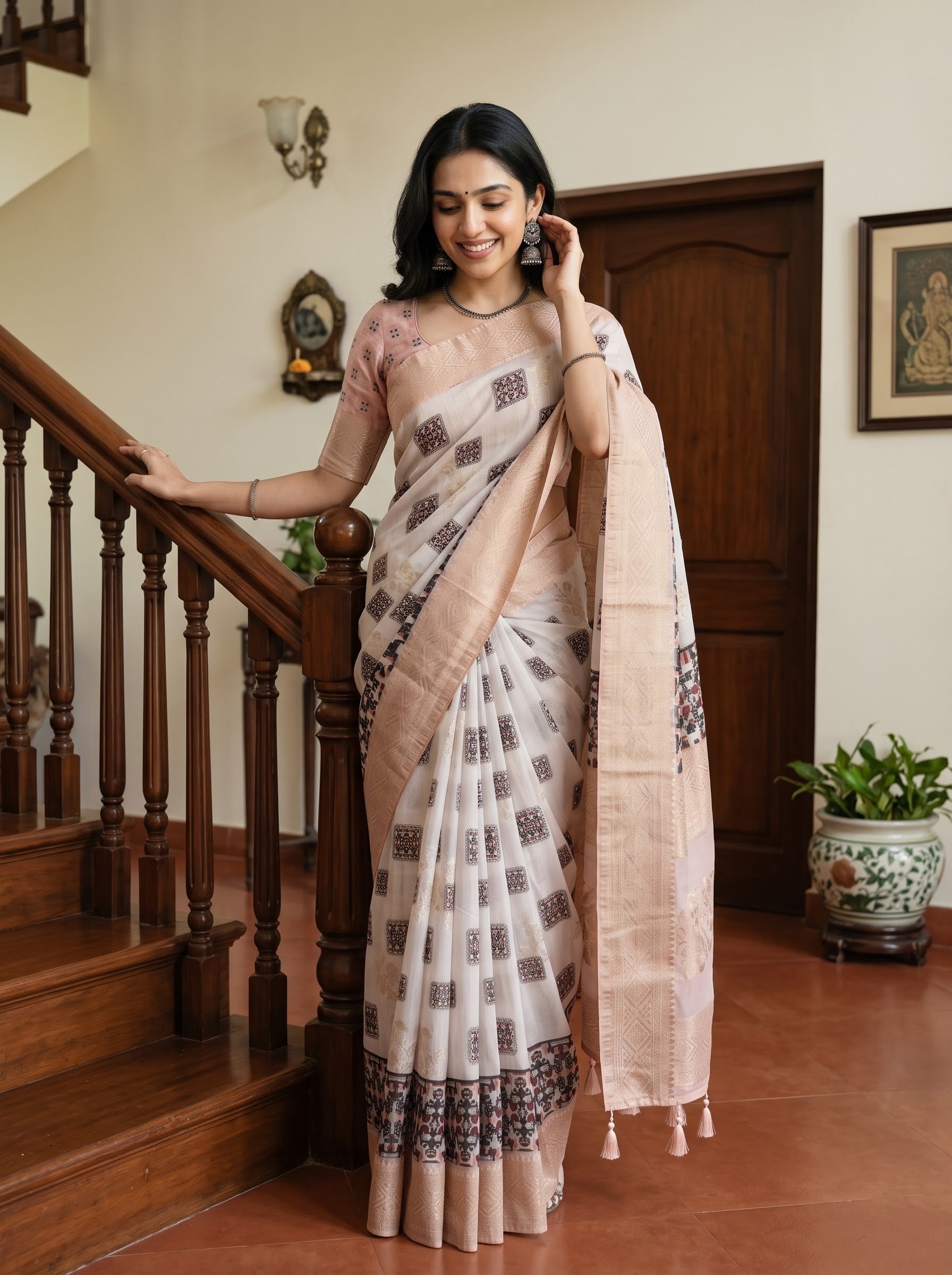 A woman wearing a traditional Indian saree stands in a home interior next to a wooden staircase. The saree has a light beige and off-white base with brown geometric block prints and a solid beige border, paired with a matching beige blouse.