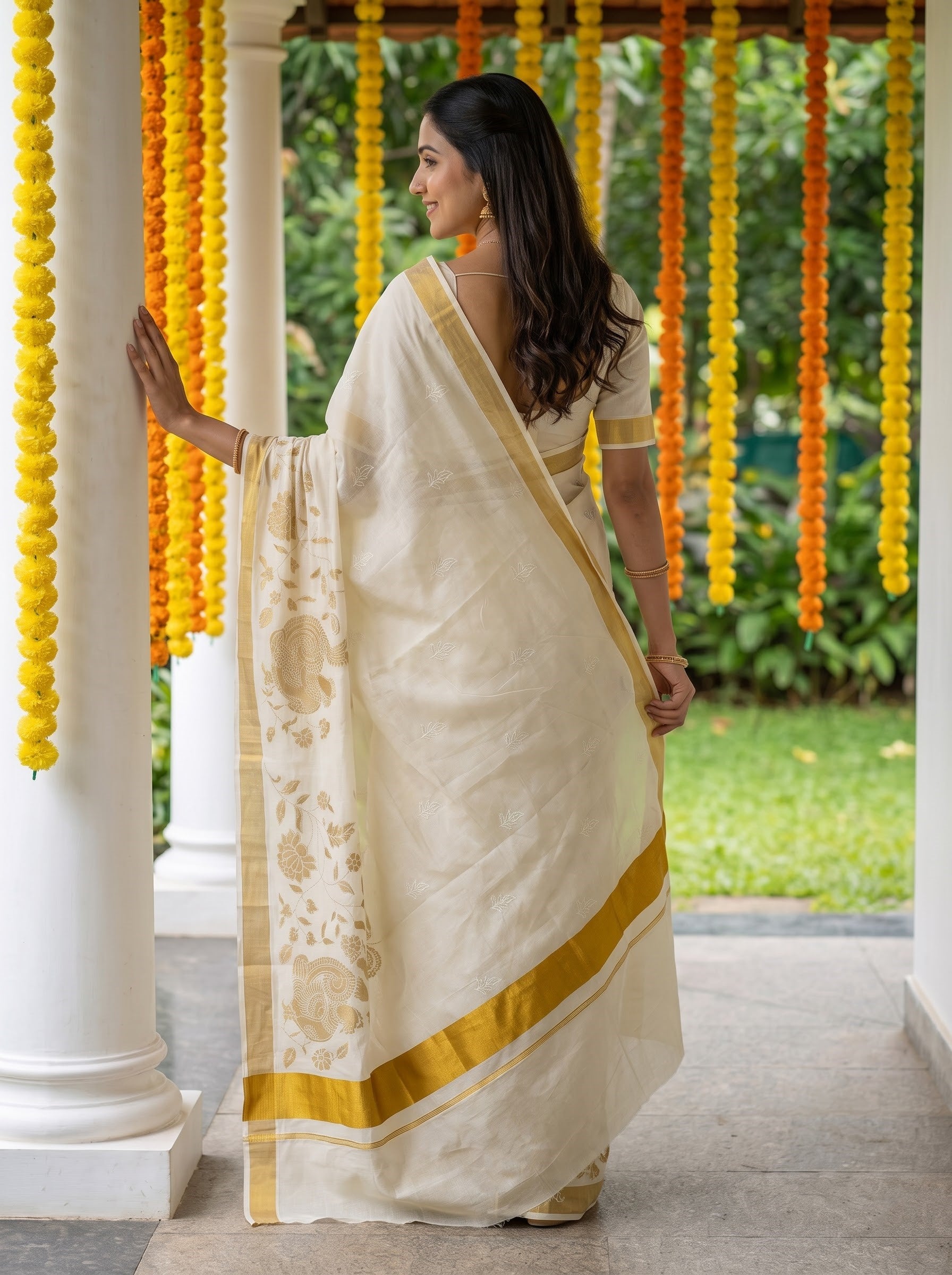 A woman wearing a cream-colored silk saree with gold border and floral motifs, standing in a garden corridor decorated with yellow marigold garlands.