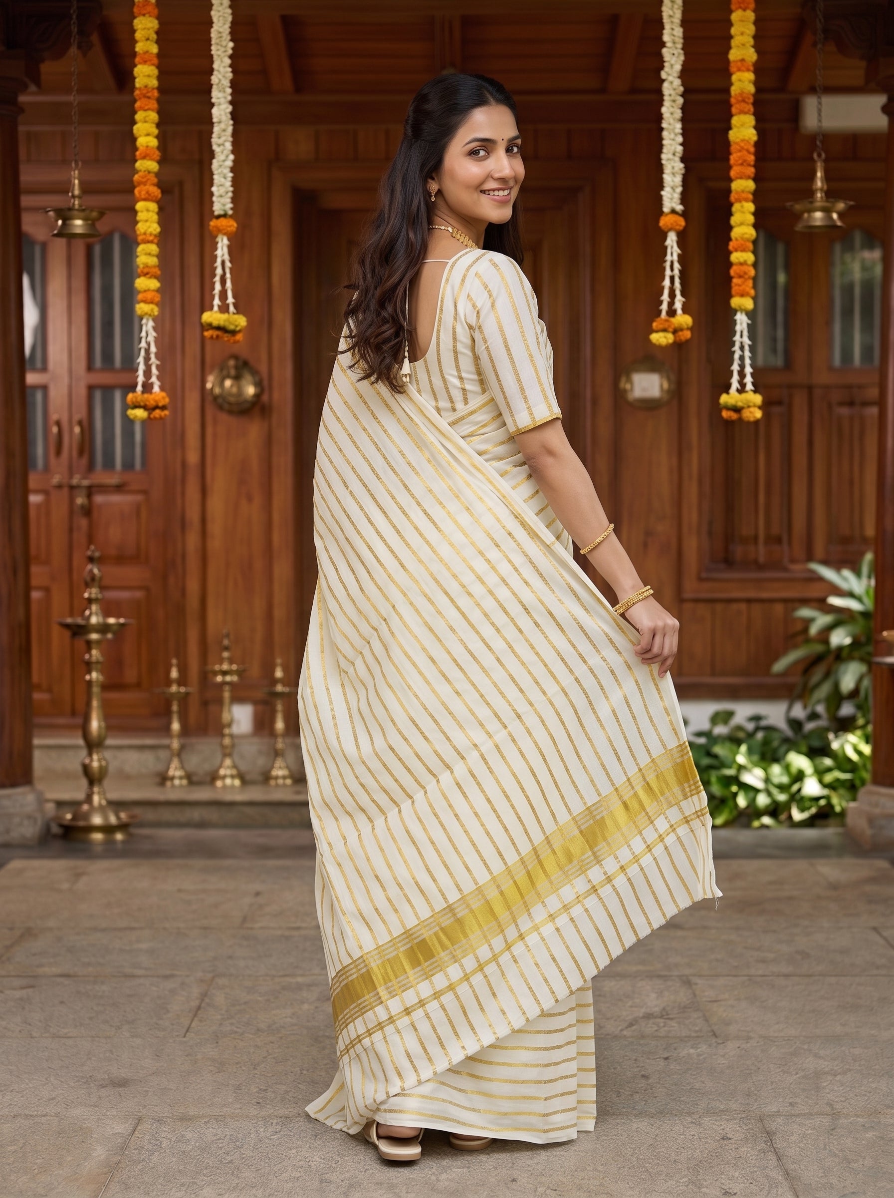 A woman wearing a cream-colored cotton saree with thin vertical gold stripes and a gold border, standing in front of a traditional wooden doorway decorated with marigold garlands.