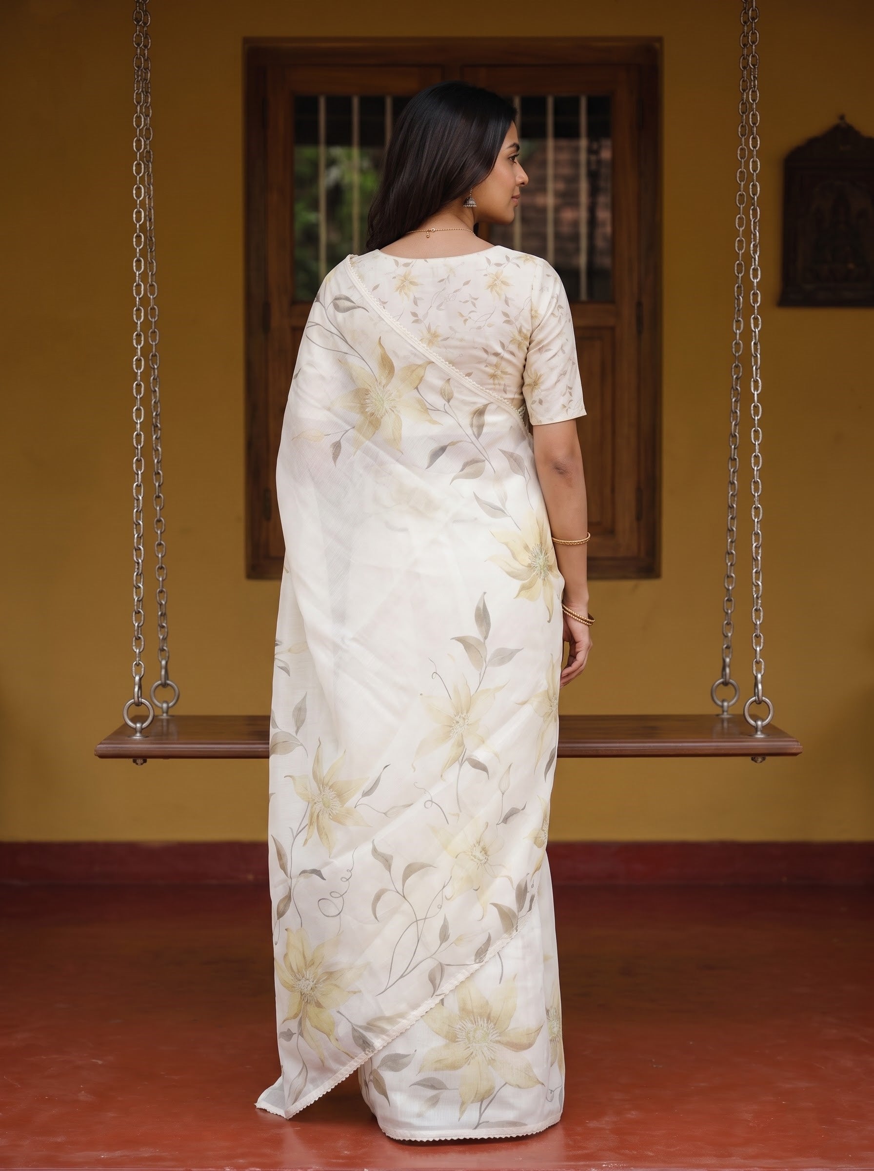 A woman stands with her back to the camera wearing a cream-colored saree with a beige and gold floral pattern, set against a rustic indoor background with a wooden swing and yellow walls.