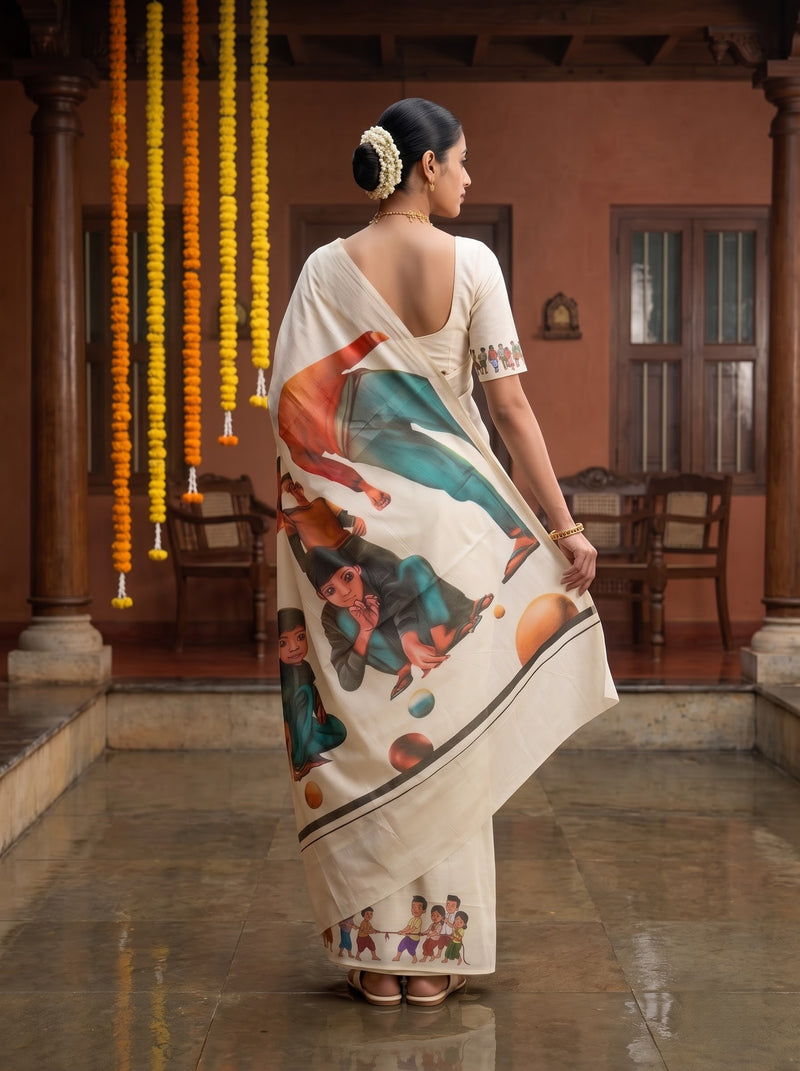 A woman stands in a courtyard wearing a cream-colored saree with colorful traditional illustrations and a white blouse, photographed from the back.