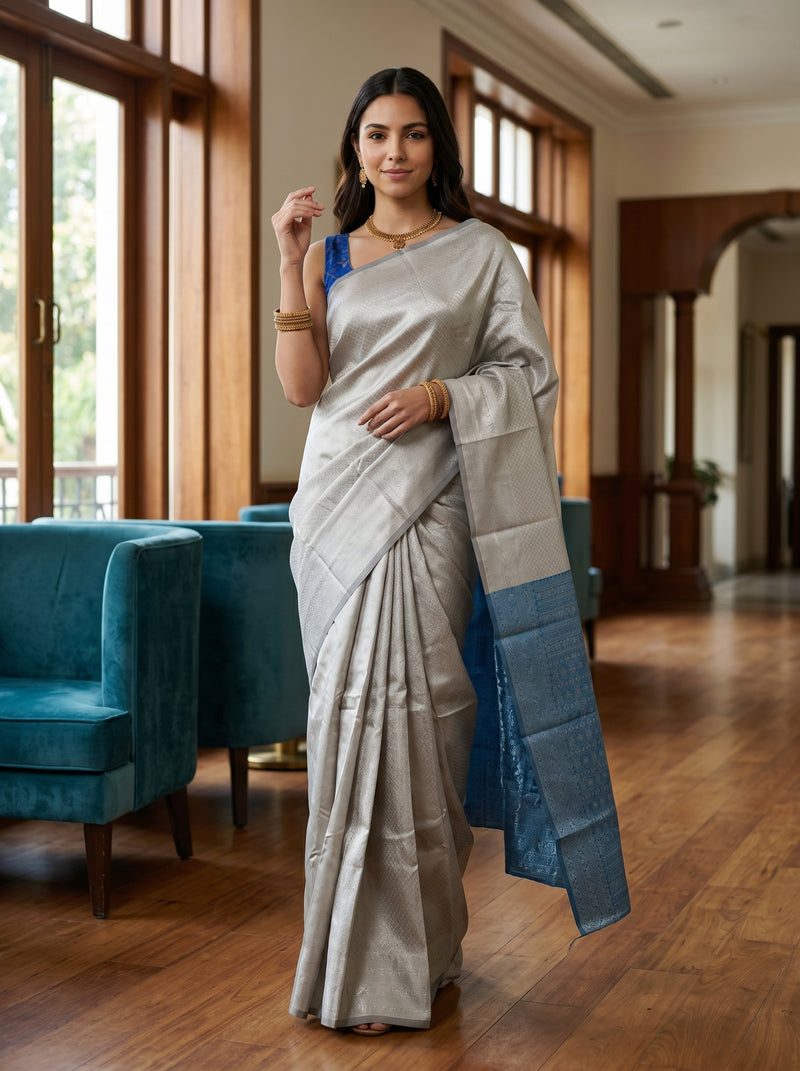 Woman in a light gray saree with blue border standing in a room with wooden floor and teal chairs.