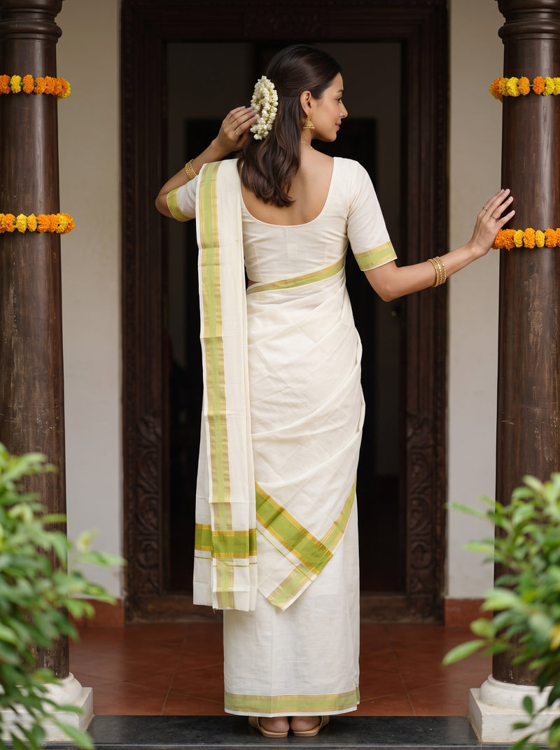 A woman wearing a white cotton saree with light green borders, standing with her back to the camera in a doorway decorated with dark pillars and yellow flower garlands.