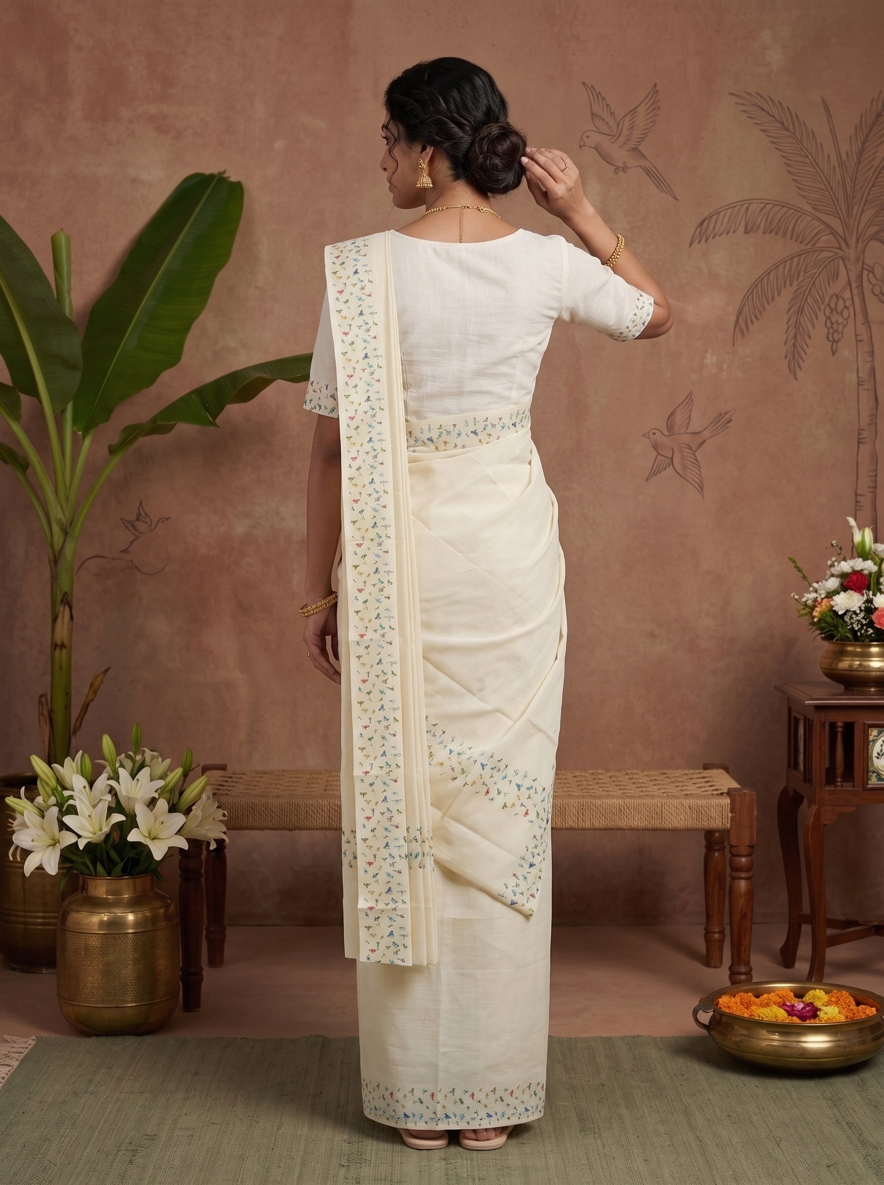 A woman stands with her back to the camera wearing a cream-colored cotton saree with a matching blouse. The saree features delicate floral embroidery in light blue and green along the border and pallu.